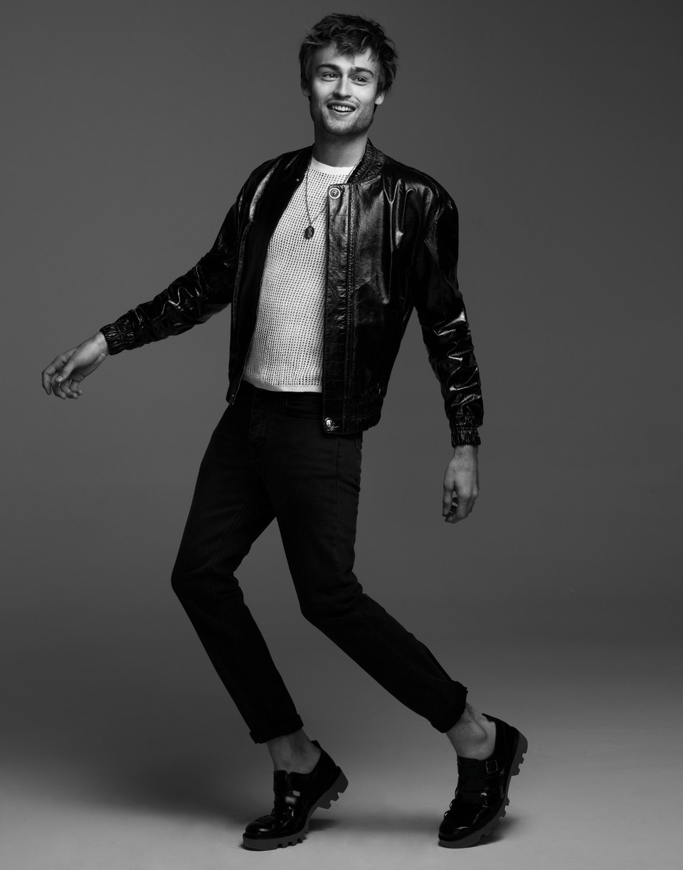 Douglas Booth stands on his tip toes while in a leather jacket and black pants